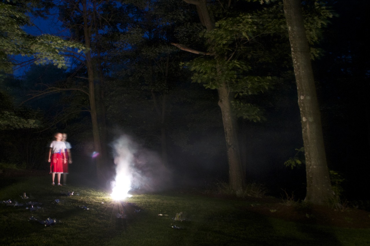 At left, a child of around 10 looks ominously on at a bright, smoking light at center-left within a dark forest. Around the
         bright light are paper wrappings: the wrappings for spent fireworks.